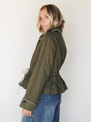 A model shot from the back-angle showing an olive utility jacket with a feminine cinched waist and cream contrast piping, paired with relaxed medium-wash denim — the kind of outfit that looks like you put in effort without trying too hard.