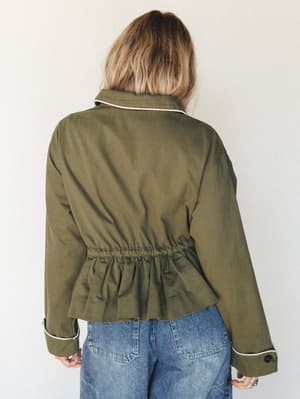 A back-view shot of a cropped olive utility jacket with a gathered peplum waist and cream piping trim, styled over wide-leg light wash jeans against a soft neutral studio wall.