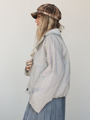A side-profile shot of a model in a cream grid-check jacket layered over a soft blue pleated skirt, topped with a brown plaid newsboy cap — the whole look has that effortless vintage-meets-weekend energy.