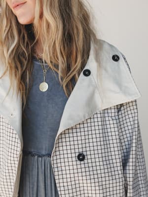 A close-up torso shot of a mixed-fabric jacket in cream and grid check layered over a washed blue dress, styled with gold layered necklaces — relaxed and intentional, like a Saturday that actually comes together.