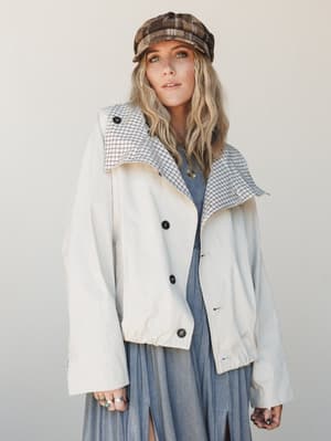 A relaxed, layered look built around a cream button-front jacket with a contrast plaid collar, worn over a dusty blue midi dress and finished with a plaid newsboy cap and stacked rings — the kind of outfit that feels vintage-inspired without trying t