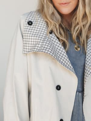 A close-up detail shot of a cream trench-style jacket with a contrast grid-check collar, layered over a washed blue top — the kind of piece that makes a simple outfit feel intentional.