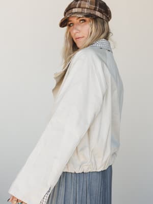A blonde model shot in three-quarter profile against a soft neutral studio wall, layering a cream bomber-style jacket over a checkered collared shirt and pleated blue-grey midi skirt, topped with a brown plaid newsboy cap — the whole look has that ef