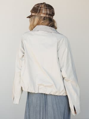 Shot from behind, a model wears a relaxed cream bomber-style jacket over a grid-check shirt and a flowy slate blue skirt, topped with a brown plaid newsboy cap — the kind of effortlessly layered look that feels like a Sunday with nowhere to be.