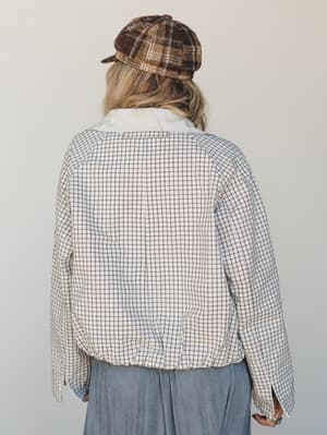 Shot from behind, she's wearing a cropped windowpane-check jacket in cream and a plaid newsboy cap in warm brown — the kind of layered, lived-in look that feels effortlessly put-together on a Tuesday.