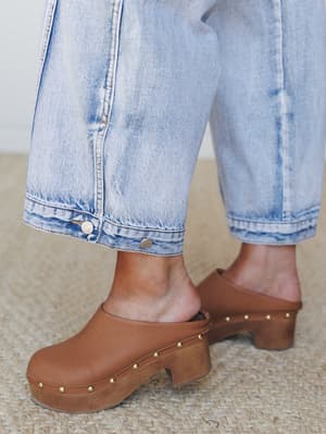 A close-up of wide-leg light wash jeans with button hem detail, styled with tan leather platform clogs — the kind of effortless pairing that looks like you've been dressing this way your whole life.