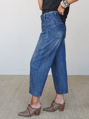 A side-angle shot of relaxed barrel-leg jeans in medium denim, styled with a faded black tee, leopard print mules, and a stacked mix of turquoise and pearl bracelets — the kind of outfit that looks effortless but was definitely thought about.