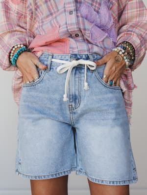 A close-up of light wash denim bermuda shorts with a white drawstring waist, styled with a pink plaid shirt and a stack of beaded and stone bracelets — the kind of effortless summer outfit that looks like it took no thought but absolutely did.