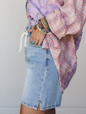 A close-cropped detail shot of light wash denim shorts paired with a pastel plaid oversized top, the hand tucked into the pocket showing off a stacked mix of beaded bracelets and stone rings — the kind of accessory moment that makes the whole outfit.