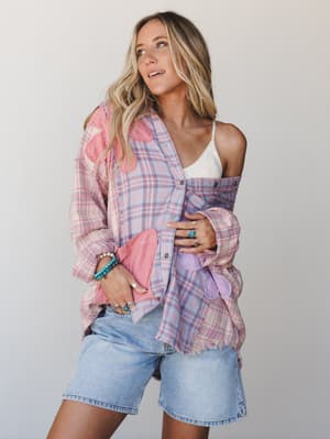 A relaxed, sun-warmed look built around light wash denim shorts layered under an oversized pink plaid flannel shirt worn open and off-shoulder — the kind of outfit that looks effortless because it actually is.
