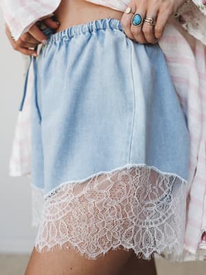 A close-up detail shot of light denim shorts with a wide scalloped lace hem, styled with a floral and gingham layered top and stacked boho rings — the kind of piece that feels equally at home on a porch or at a summer market.