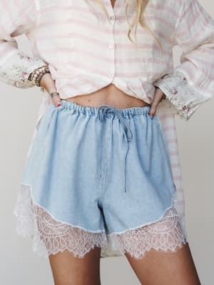 A close mid-shot of light wash denim pull-on shorts with a scalloped lace hem, styled with a pink striped linen button-down tied at the waist and stacked boho bracelets — soft, romantic, and very much the vibe of a slow morning that somehow looks thi