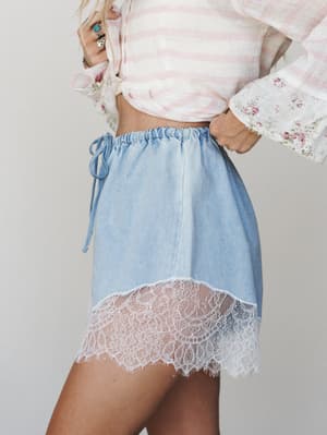 A soft, dreamy close-up of light denim mini shorts with a scalloped white lace hem, styled with a pink striped crop top with floral cuff detail — the kind of outfit that feels like a vintage postcard come to life.