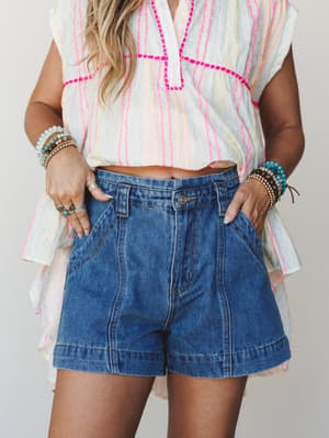 A cropped mid-shot of a model in high-waisted medium denim shorts paired with a breezy striped top with hot pink embroidery stitching — stacked boho bracelets and silver rings round out that effortless summer-ready look.