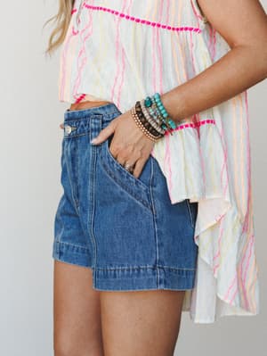 A cropped mid-shot of a breezy summer outfit — denim shorts paired with a flowy striped top and a stacked bracelet arm party that does all the talking.