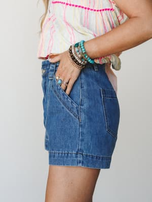 A side-angle mid-shot showing medium wash denim shorts paired with a multicolor striped top and a stacked bracelet arm party — casual, colorful, and the kind of outfit that makes a Tuesday feel like a road trip.