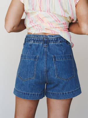 A close back-view shot showing relaxed medium-wash denim shorts with oversized patch pockets, paired with a knotted multicolor striped top — casual summer dressing that feels easy and a little nostalgic.