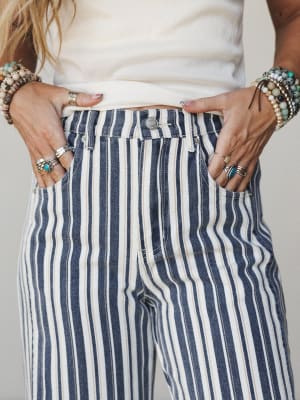 A close-up of navy and cream vertical stripe denim pants styled with a cream top and layered with stacked stone bracelets and silver rings — the kind of detail shot that makes you notice the accessories as much as the pants.
