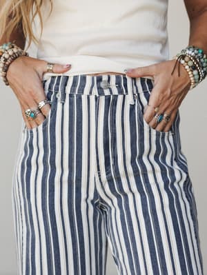 A close-up of navy and cream vertical stripe denim pants styled with a cream top and layered with stacked stone bracelets and silver rings — the kind of detail shot that makes you notice the accessories as much as the pants.