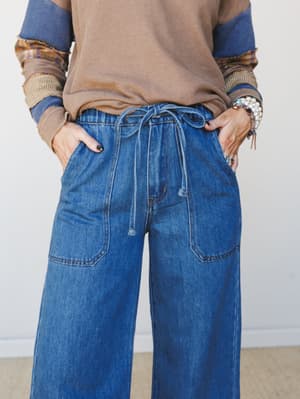 A close-up mid-shot of wide-leg denim jeans with a relaxed drawstring waist and patch pockets, styled with a layered mixed-texture top and stacked bracelets for an effortlessly lived-in look.