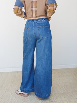 A back-view shot of wide-leg pull-on jeans in medium blue denim, styled with a patchwork plaid cropped top and leopard sandals against a soft studio backdrop — relaxed and effortlessly put-together.