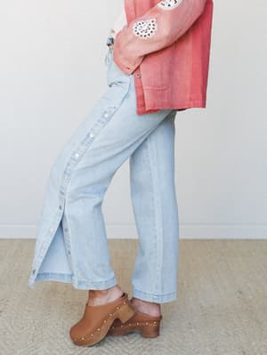 A side-angle detail shot of wide-leg light wash jeans with a side snap button placket, styled with a rosy washed jacket with crochet appliqué and brown studded clogs — easy, vintage-leaning, and very much the outfit you'd wear to feel like yourself o