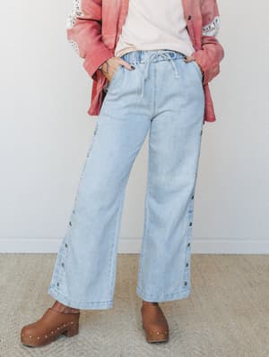 Light wash wide-leg denim pants with side snap buttons and a drawstring elastic waist, styled with a rose-toned linen jacket with crochet trim and brown wooden clogs — casual, expressive, and very much the kind of outfit you wear when you want to fee