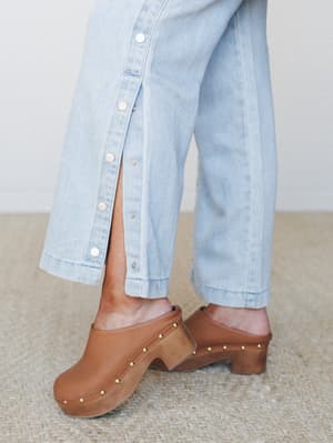 A close-up of wide-leg light denim jeans with a snap-button side slit, styled with tan studded clogs on a woven rug — the kind of detail shot that makes you immediately want to know where the shoes are from.