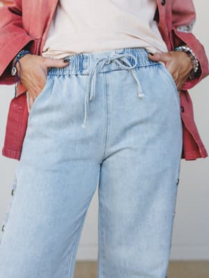 A close-up mid-shot of light stonewash wide-leg jeans with a relaxed elastic drawstring waist and side snap buttons, layered under a dusty rose shacket — the kind of outfit that looks effortless but clearly wasn't an accident.