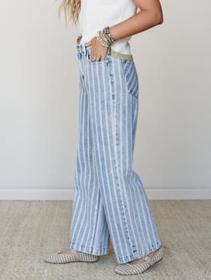 A side-profile shot of wide-leg striped denim jeans styled with a white textured top, woven flats, and layered stone bracelets — relaxed and collected with a quiet vintage feel.