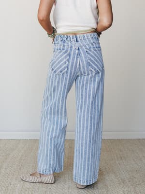 A back-view shot of wide-leg light denim jeans with a vertical pinstripe print and elastic waistband, styled with a cropped cream top, stacked bead bracelets, and woven flats — easy and expressive, like a Saturday that actually came together.