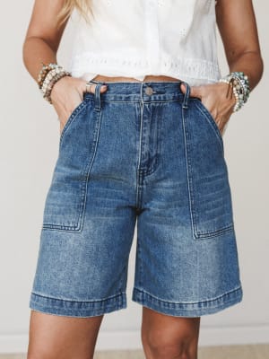 A cropped torso shot of medium-wash denim Bermuda shorts styled with a white eyelet top and stacked beaded bracelets — the kind of effortless summer outfit that works just as hard at the farmers market as it does on a weekend road trip.