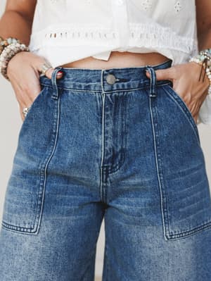 A close-up shot of high-waisted wide-leg denim paired with a white eyelet crop top — hands tucked into patch pockets, stacked bracelets on both wrists, the kind of effortless outfit you reach for on repeat.