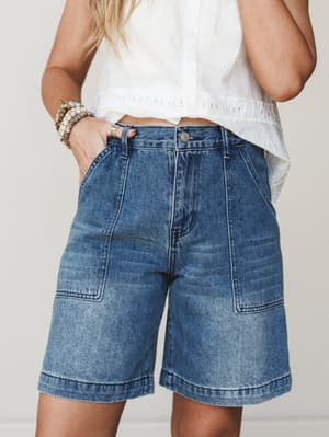 A cropped mid-shot of relaxed, longer-length denim shorts paired with a white eyelet top — the kind of effortless summer outfit that goes from the farmers market straight to lunch.