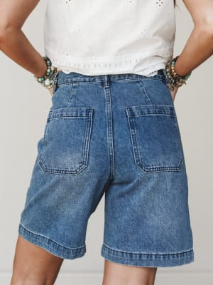 A back-view shot of relaxed denim bermuda shorts paired with a white eyelet crop top and layered beaded bracelets — casual, expressive, and the kind of outfit that looks effortless on a summer errand run.