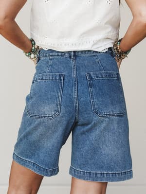 A back-view shot of relaxed denim bermuda shorts paired with a white eyelet crop top and layered beaded bracelets — casual, expressive, and the kind of outfit that looks effortless on a summer errand run.