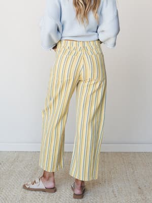 Shot from behind, a model pairs buttery yellow striped barrel pants with a soft blue knit sweater and neutral slide sandals — a relaxed, sun-washed combo that feels like a slow Saturday morning.