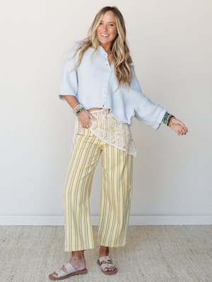 A smiling model pairs butter-yellow striped wide-leg pants — accented with a cream floral lace waistband panel — with a powder blue cropped knit cardigan and white buckle sandals, the whole look feeling like a breezy spring afternoon with nowhere to