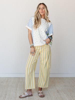 A sun-warmed mix-print moment — yellow and blue striped wide-leg pants paired with a patchwork floral and stripe tee, sandals, and a stack of bracelets that looks like it grew there naturally.