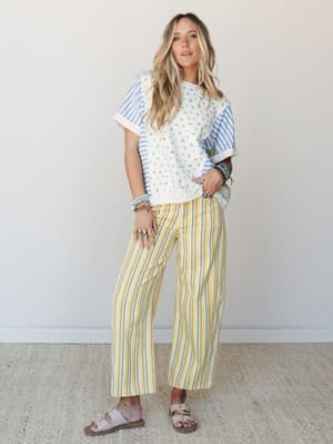 A sun-warmed mix-print moment — yellow and blue striped wide-leg pants paired with a patchwork floral and stripe tee, sandals, and a stack of bracelets that looks like it grew there naturally.