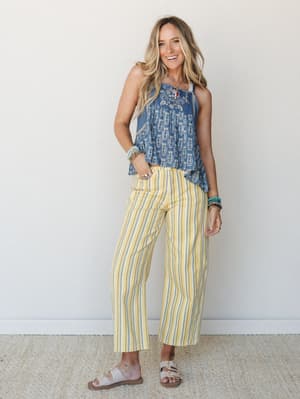 A smiling model pairs breezy yellow striped barrel pants with a blue printed sleeveless top, stacked boho bracelets, and flat sandals — the kind of effortless summer outfit that looks like it happened on purpose.