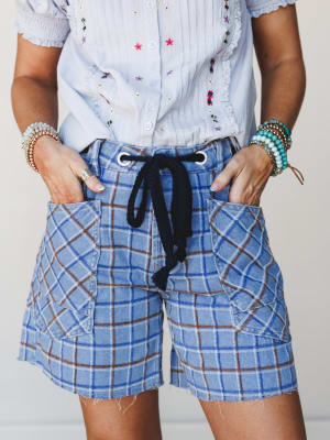 A close-up mid-shot of plaid denim shorts with a rope drawstring and oversized patch pockets, styled with a floral embroidered button-up and stacked boho bracelets — casual and collected with just enough personality.