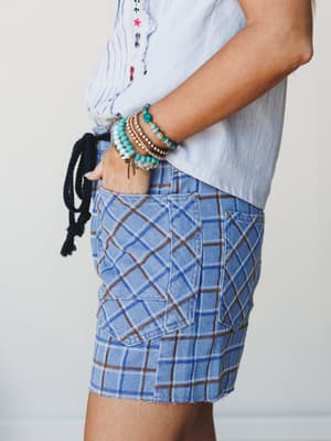 A close side-crop of plaid denim shorts with patch pockets and raw-hem edges, paired with a light blue embroidered top and a stack of turquoise and gold beaded bracelets — casual and collected with just enough personality.