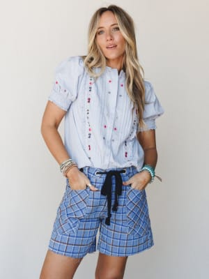 A light and layered summer look — embroidered blouse tucked loosely into plaid denim shorts with a drawstring tie, styled with stacked bracelets for that effortless boho-casual feel.