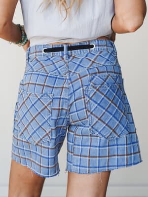 A back-view close-up of plaid denim shorts with patchwork-style paneling and a raw hem, styled with a white tee and turquoise stacked bracelets — the kind of shorts that feel vintage-found but totally intentional.