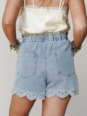 A back-view shot of light-wash denim shorts with eyelet scallop trim at the hem and waist, styled with a cream lace-trim cami and stacked boho bracelets — the kind of outfit that looks just as good on a patio as it does at a Sunday market.