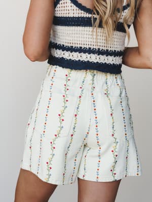 A back-view shot of cream denim shorts covered in delicate vertical floral vine embroidery, paired with a navy and cream striped crochet crop top — the kind of outfit that makes a Saturday feel intentional.