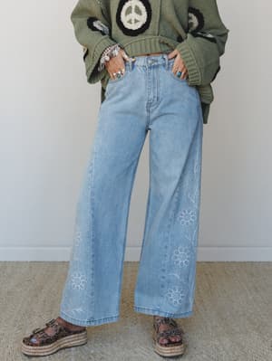 Light wash wide-leg jeans with tonal floral embroidery along the hem, styled with a cropped olive green peace-sign crochet sweater, stacked silver rings and bracelets, and platform espadrille sandals — the whole look reads like a 70s record collectio