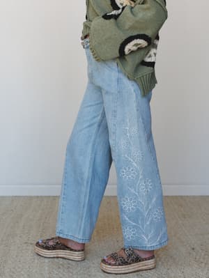 A cropped side-angle shot of wide-leg light wash jeans with tone-on-tone floral vine embroidery running up the leg, styled with a chunky patterned knit sweater and leopard print platform sandals — casual and expressive in the best way.