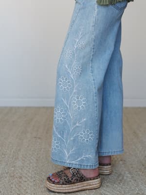 A close detail shot of wide-leg light wash denim jeans with delicate white daisy embroidery climbing up the leg, styled with leopard print platform sandals on a woven rug — casual boho with a little personality.